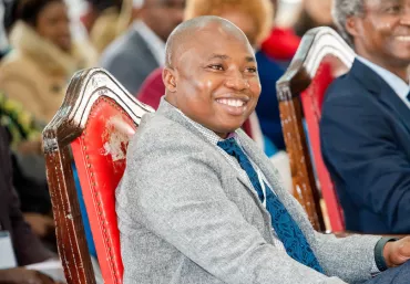 Jerry Ouner, dressed in a suit and sitting on an elaborate red chair smiling while listening to a speaker.